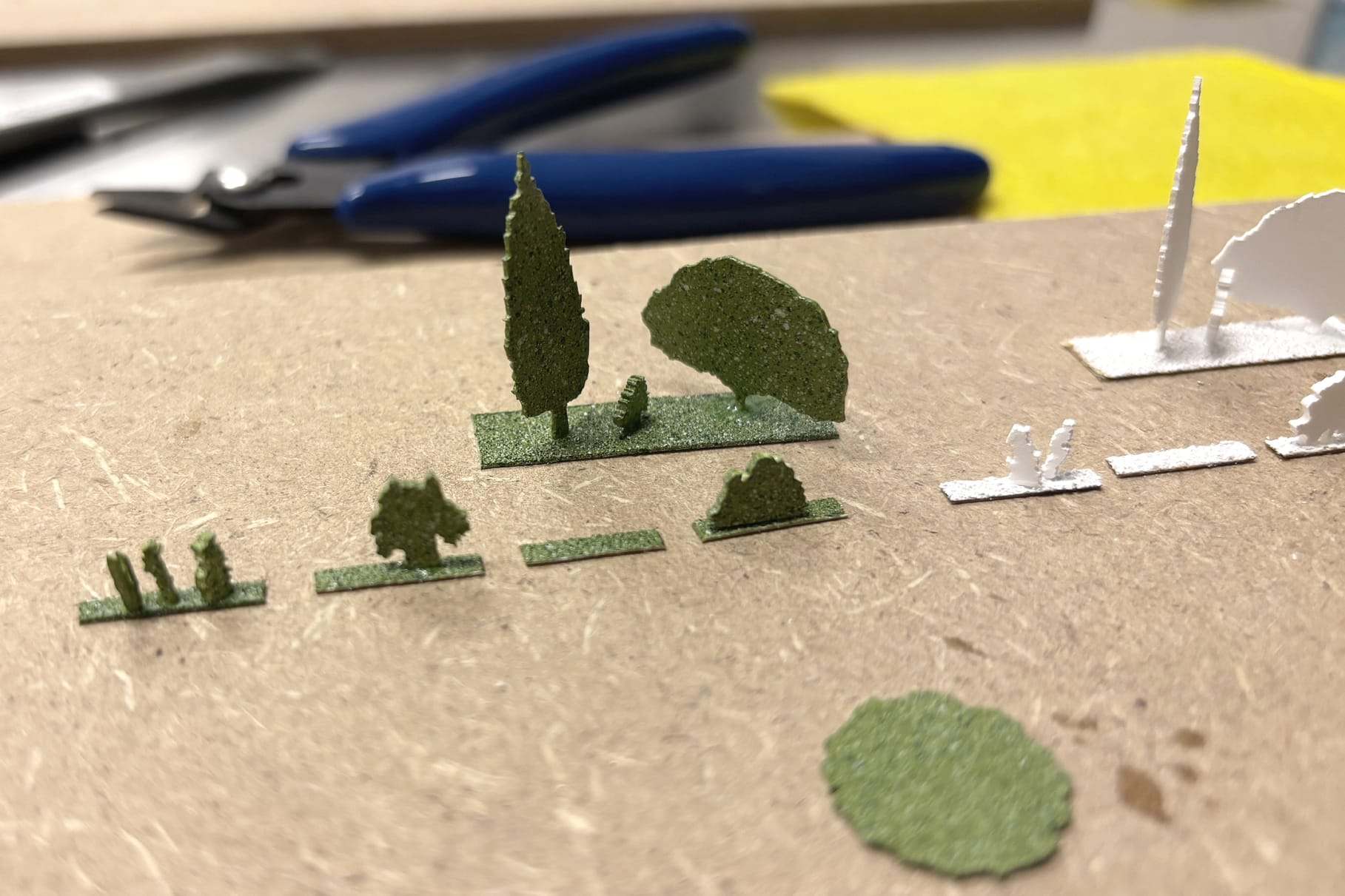 Small green model trees and white pieces on a work surface with pliers