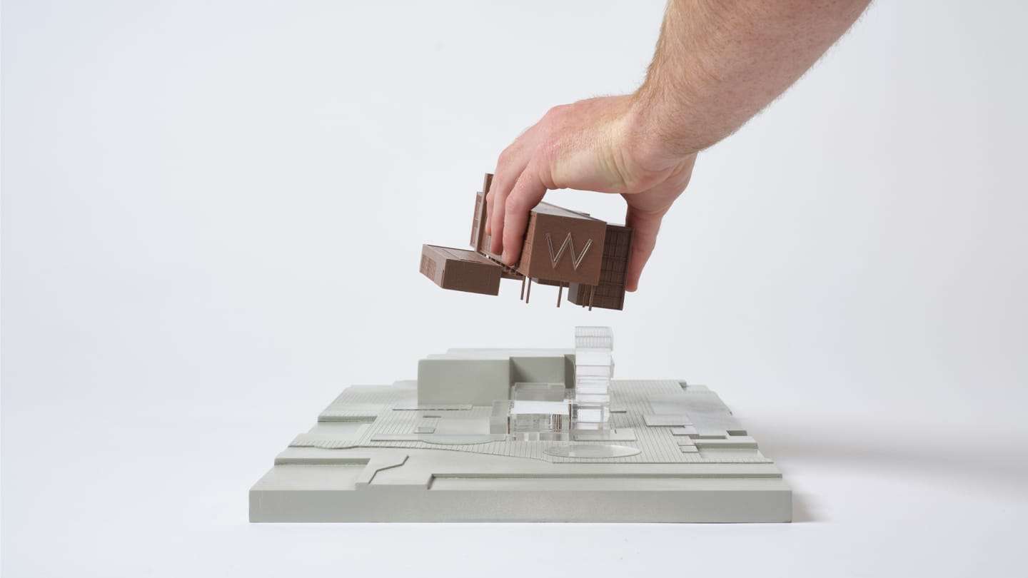 A hand lifts the pre-renovation building off the site base of the 1:200 model
