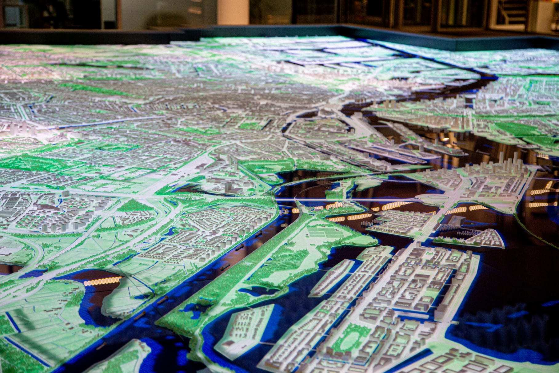 The city model lit from below in blue and green, showing the waterways across Amsterdam